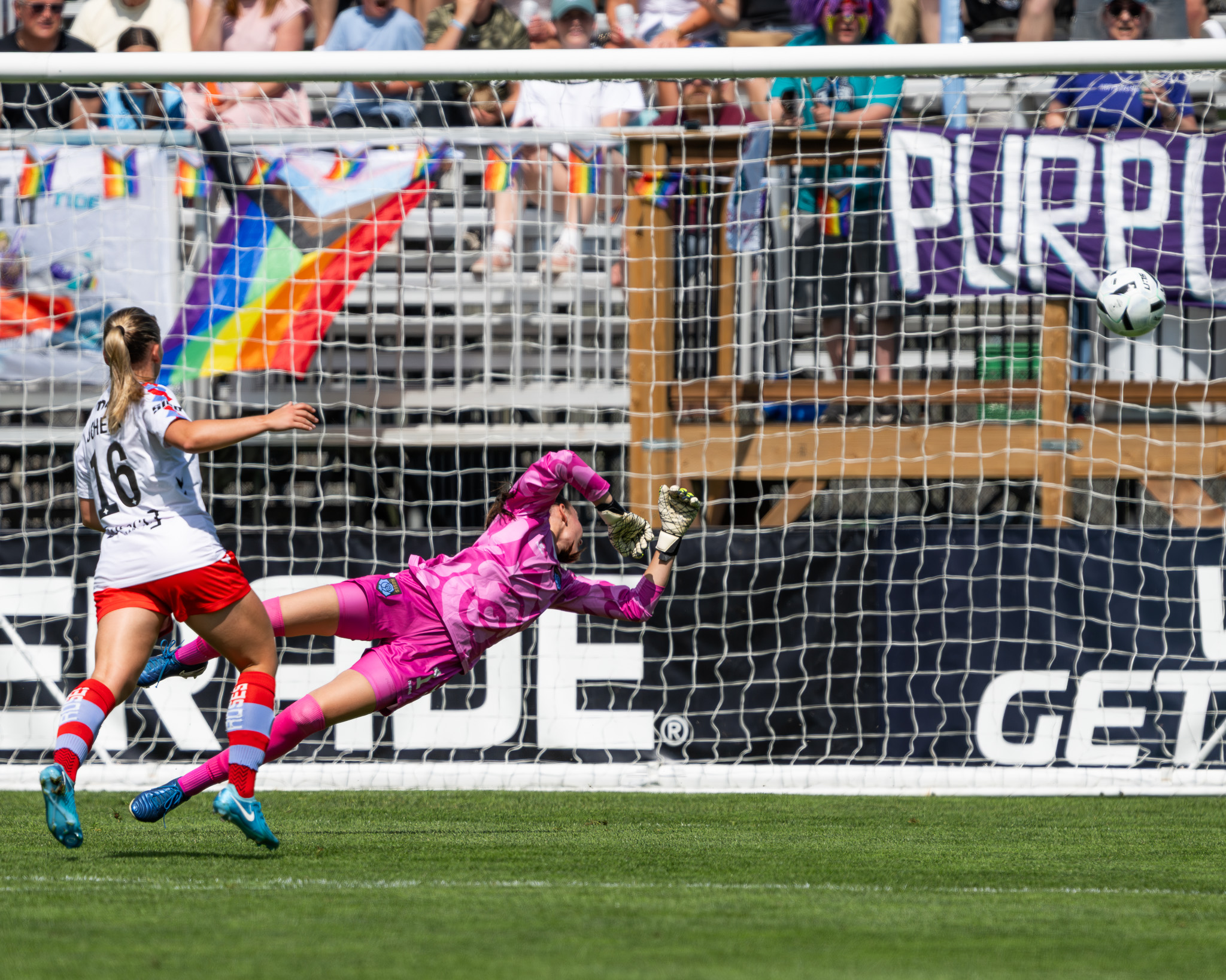 Goalkeeper in a bright pink kit dives full extension toward the top corner as a shot heads toward the net, with a defender trailing and fans in the background.